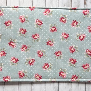 May include: A rectangular placemat with a light blue background and a pattern of red and pink flowers with green leaves. The background has small white polka dots. The edges are neatly stitched.