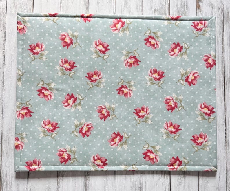 May include: A rectangular placemat with a light blue background and a pattern of red and pink flowers with green leaves. The background has small white polka dots. The edges are neatly stitched.
