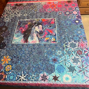 May include: A colorful quilt featuring a central image of a white horse with a flowing mane, surrounded by vibrant blues, pinks, and purples. The quilt has a detailed pattern of floral and geometric shapes, with a pink border.
