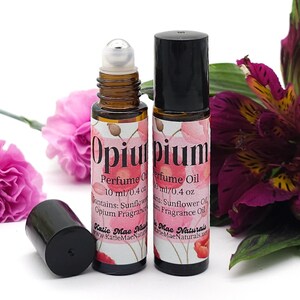 Puede incluir: Dos frascos de vidrio &aacute;mbar de 10 ml / 0,4 oz con tapas negras y bolas de rodillo plateadas. Los frascos est&aacute;n etiquetados con "Opium Perfume Oil" con un dise&ntilde;o floral de acuarela. El texto "Contiene: Aceite de girasol, Aceite de fragancia de opio" est&aacute; impreso en la etiqueta. Los frascos est&aacute;n sobre una superficie blanca con flores rosas y moradas.
