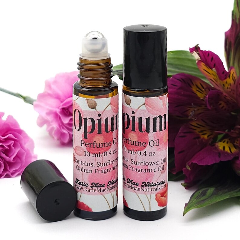 Puede incluir: Dos frascos de vidrio &aacute;mbar de 10 ml / 0,4 oz con tapas negras y bolas de rodillo plateadas. Los frascos est&aacute;n etiquetados con "Opium Perfume Oil" con un dise&ntilde;o floral de acuarela. El texto "Contiene: Aceite de girasol, Aceite de fragancia de opio" est&aacute; impreso en la etiqueta. Los frascos est&aacute;n sobre una superficie blanca con flores rosas y moradas.
