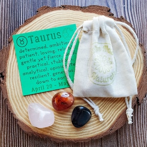 May include: A set of three crystals in a small white drawstring bag with a green tag that reads "Taurus" and lists the astrological sign's traits. The crystals are pink, orange, and black.