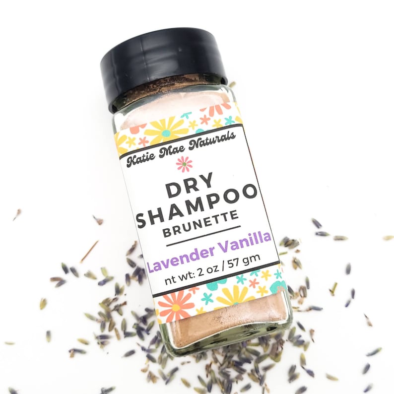 May include: A small glass jar with a black lid filled with a light brown powder. The label is white with a floral design and the text "Katie Mae Naturals Dry Shampoo Brunette Lavender Vanilla nt wt: 2 oz/57 gm".