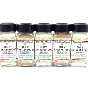 May include: Six small glass jars with black lids containing dry shampoo powder. Each jar has a colorful label with the text "Katie Mae Naturals Dry Shampoo" and a different color and scent: Pink Grapefruit Blonde, Unscented Brunette, Clary Sage Lime Blonde, Orange Jasmine Brunette, Vanilla Bean Blonde.