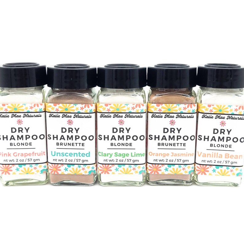 May include: Six small glass jars with black lids containing dry shampoo powder. Each jar has a colorful label with the text "Katie Mae Naturals Dry Shampoo" and a different color and scent: Pink Grapefruit Blonde, Unscented Brunette, Clary Sage Lime Blonde, Orange Jasmine Brunette, Vanilla Bean Blonde.