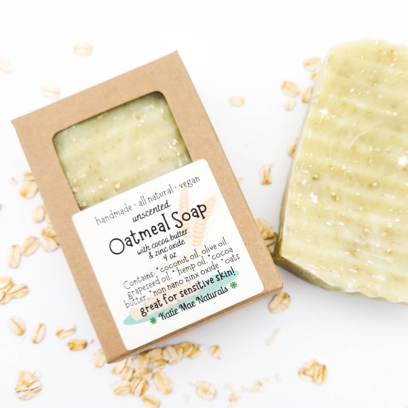May include: A brown cardboard box with a clear window showing a light green bar of soap. The box has a white label with the text "handmade all natural. vegan unscented Oatmeal Soap with cocoa butter & zinc oxide 4 oz Contains: "coconut oil, olive oil. grapeseed oil. hemp oil. cocoa butter, "non nano zinx oxide. "oats great for sensitive skin! *Katie Mae Naturals *"