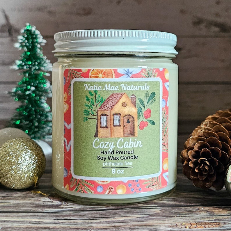 Puede incluir: Una vela en tarro de vidrio con tapa blanca y una etiqueta que dice "Cozy Cabin". La etiqueta presenta una peque&ntilde;a ilustraci&oacute;n de una casa y el texto "Katie Mae Naturals". La vela es de 255 gramos y est&aacute; rodeada de decoraciones navide&ntilde;as.