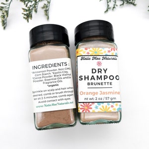 May include: Two small glass jars with brown powder inside. The jar on the left has a label that reads "INGREDIENTS: "Arrowroot Powder. Non GMO Corn Starch. "Kaolin Clay. "Cocoa Powder, Black Walnut Powder, Essential Oils and/or Fragrance Oils "organic Sprinkle on scalp and hair where desired, comb or brush through let sit 2-3 minutes, comb again Avoid contact with eyes. www.Katie Mae Naturals.net". The jar on the right has a label that reads "Katie Mae Naturals DRY SHAMPOO BRUNETTE Orange Jasmine nt wt: 2 oz / 57 gm".