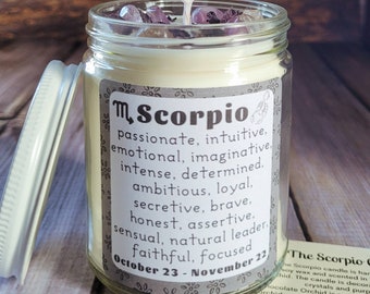 Puede incluir: Una vela de vidrio en un frasco con una tapa blanca y una etiqueta que dice "Scorpio" con el símbolo astrológico y las fechas "23 de octubre - 22 de noviembre". La etiqueta también enumera los rasgos de personalidad asociados con el signo zodiacal Escorpio. La vela está coronada con cristales morados y transparentes.