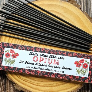 May include: Black incense sticks with gold tips are arranged on a wooden surface. A box of "Opium" incense sticks is visible, with the text "Katie Mae Naturals" and "20 Hand Dipped Incense Sticks". The box features red poppy illustrations and a decorative floral border.