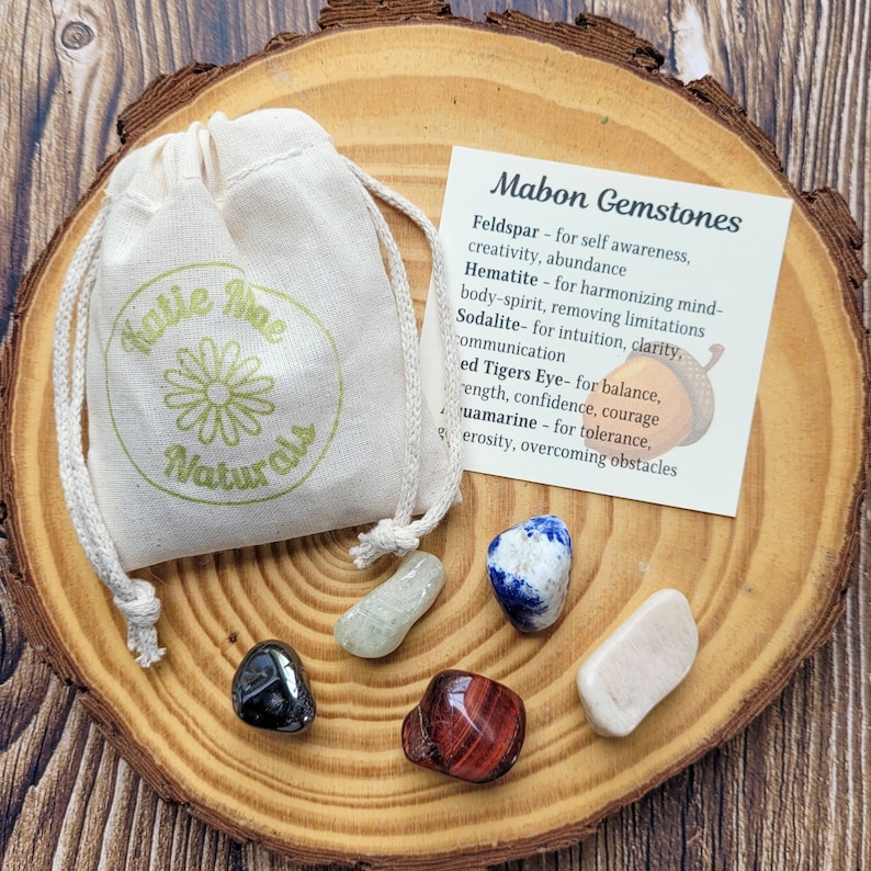 May include: A set of six gemstones in a small white drawstring bag on a wooden surface. The gemstones are: feldspar, hematite, sodalite, tigers eye, aquamarine, and a white stone. A card with the text "Mabon Gemstones" and a list of the stones and their properties is next to the bag.