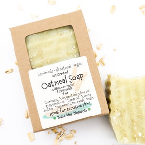 May include: A brown cardboard box with a clear window showing a bar of oatmeal soap. The box has a white label with the text "handmade - all natural - vegan unscented Oatmeal Soap with cocoa butter & zinc oxide 4 oz Contains: coconut oil, olive oil, grapeseed oil, hemp oil, cocoa butter, non nano zinc oxide, oats great for sensitive skin! *Katie Mae Naturals*"