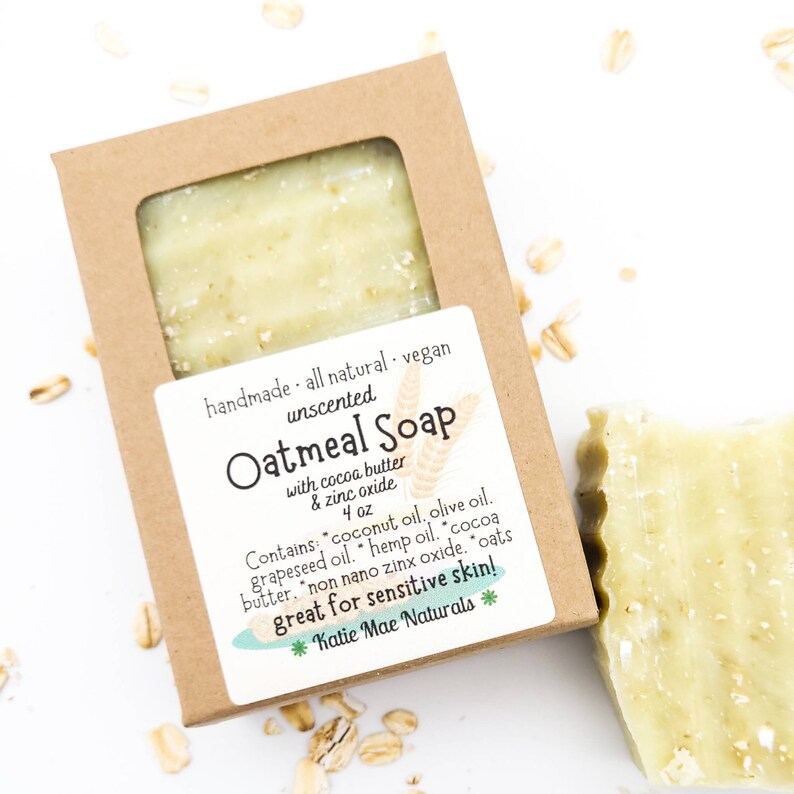 May include: A brown cardboard box with a clear window showing a bar of oatmeal soap. The box has a white label with the text "handmade - all natural - vegan unscented Oatmeal Soap with cocoa butter & zinc oxide 4 oz Contains: coconut oil, olive oil, grapeseed oil, hemp oil, cocoa butter, non nano zinc oxide, oats great for sensitive skin! *Katie Mae Naturals*"