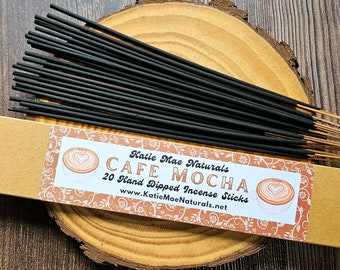 Puede incluir: Una caja de 20 varillas de incienso sumergidas a mano con un fondo marrón y una etiqueta que dice "Katie Mae Naturals Cafe Mocha 20 Hand Dipped Incense Sticks www.KatieMaeNaturals.net". La etiqueta tiene un diseño floral y una imagen de una taza de café con un corazón en la espuma.