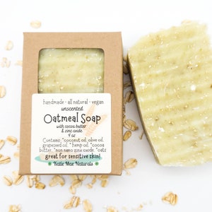 May include: A brown cardboard box with a clear window showing a bar of oatmeal soap. The label reads "handmade all natural vegan unscented Oatmeal Soap with cocoa butter & zinc oxide 4 oz Contains: coconut oil, olive oil, grapeseed oil, hemp oil, cocoa butter, non nano zinc oxide, oats great for sensitive skin! *Katie Mae Naturals*"