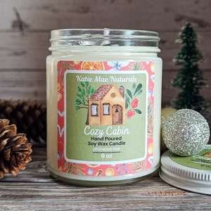 Puede incluir: Una vela en tarro de cristal con tapa. La etiqueta dice "Cozy Cabin" con un dibujo de una casa. La vela es de cera de soja vertida a mano y sin ftalatos. El tarro es de 255 g.