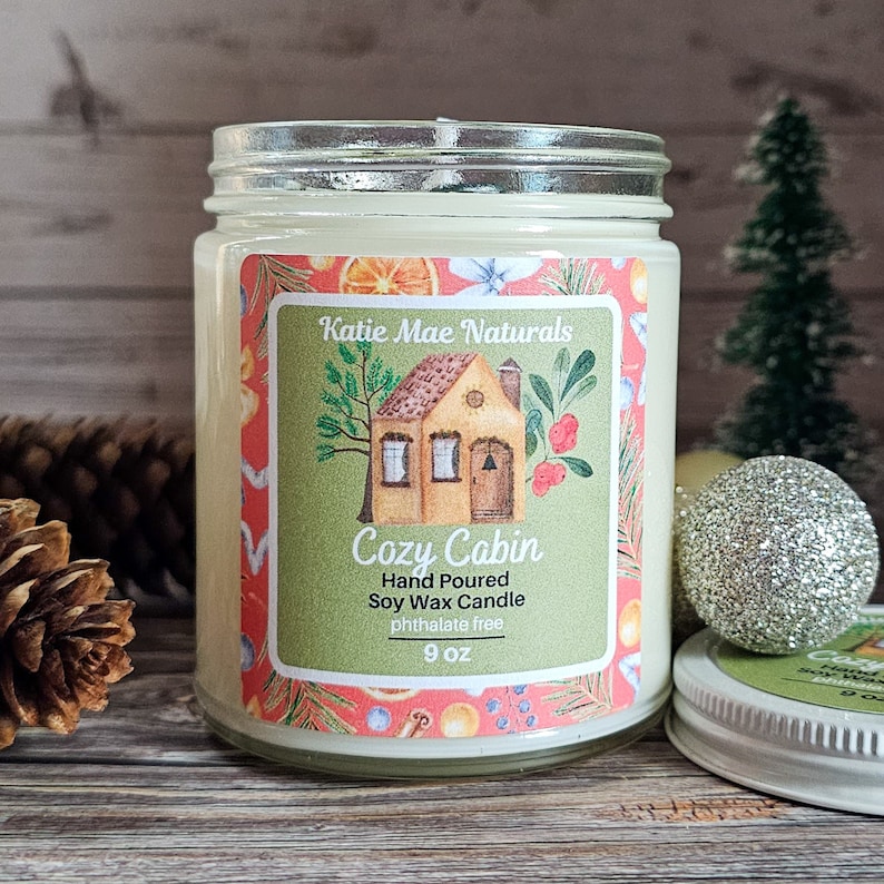 Puede incluir: Una vela en tarro de cristal con tapa. La etiqueta dice "Cozy Cabin" con un dibujo de una casa. La vela es de cera de soja vertida a mano y sin ftalatos. El tarro es de 255 g.