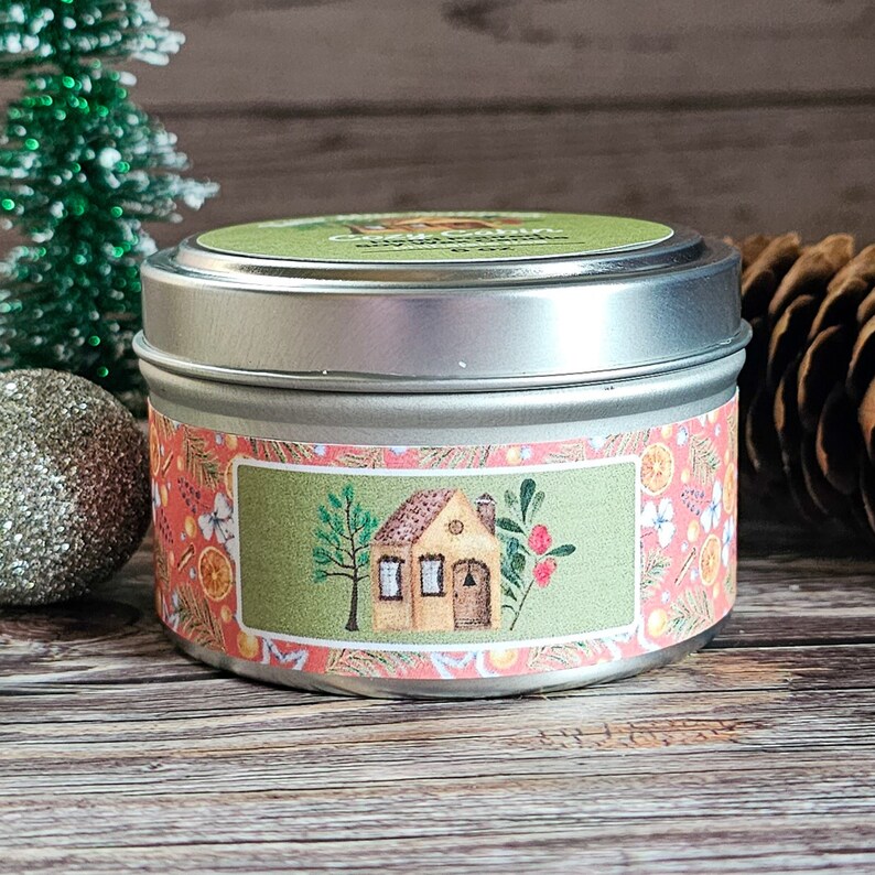 May include: A silver tin candle with a festive design. The label features a small house with a red door, a green tree, and red berries. The background is a pink and orange pattern with oranges and butterflies.