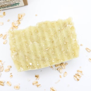 May include: A close-up of a light green bar of soap with oatmeal flakes. The soap is on a white surface with scattered oatmeal flakes.