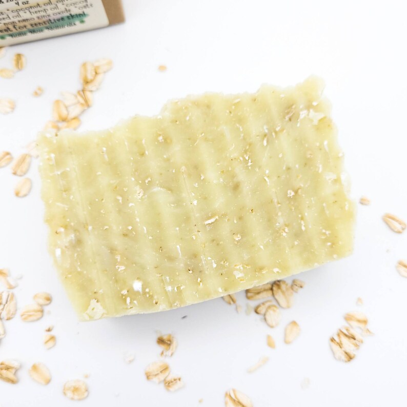 May include: A close-up of a light green bar of soap with oatmeal flakes. The soap is on a white surface with scattered oatmeal flakes.