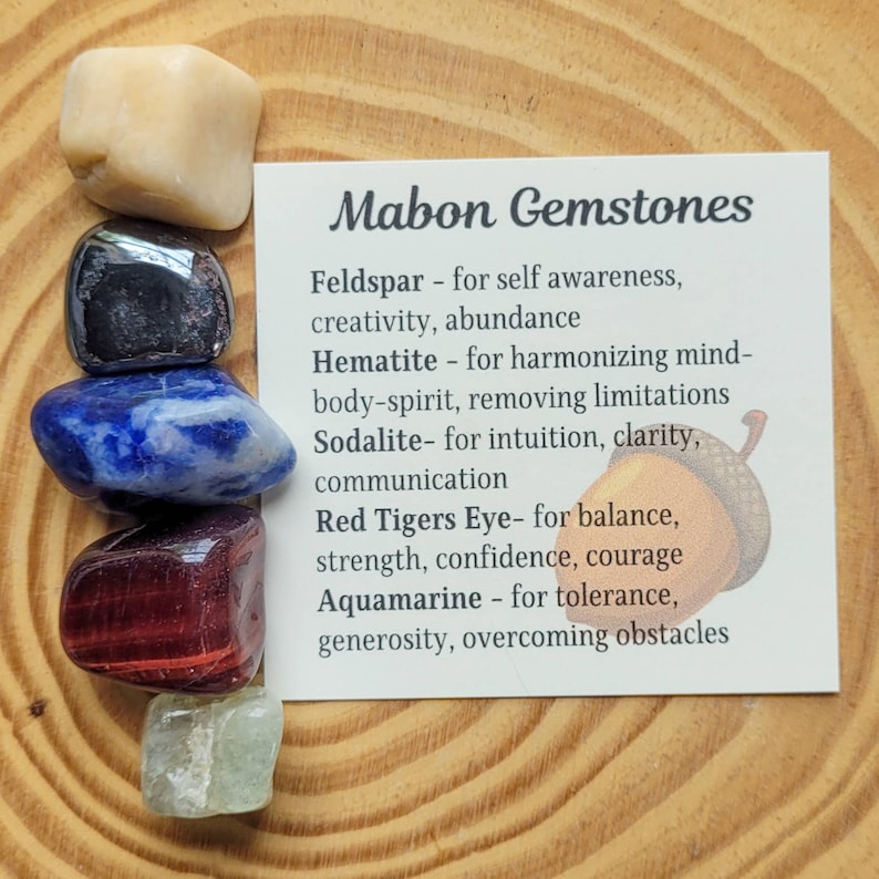 May include: A set of seven gemstones, including feldspar, hematite, sodalite, red tiger's eye, and aquamarine, arranged in a circle on a wooden surface. The gemstones are labeled with their names and associated properties, such as self-awareness, creativity, abundance, harmonizing mind-body-spirit, removing limitations, intuition, clarity, communication, balance, strength, confidence, courage, tolerance, generosity, and overcoming obstacles. Mabon Gemstones.