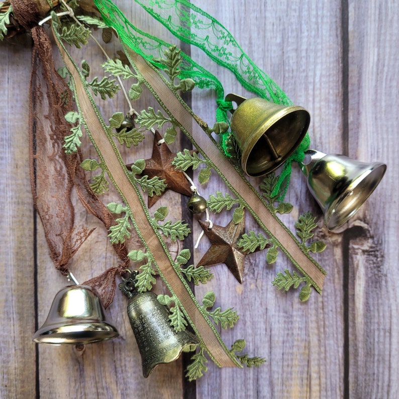 Pu&ograve; includere: Un set di quattro campane dorate con accenti di nastro e pizzo verde e marrone. Le campane sono legate insieme con un nastro marrone e hanno un aspetto rustico e vintage.