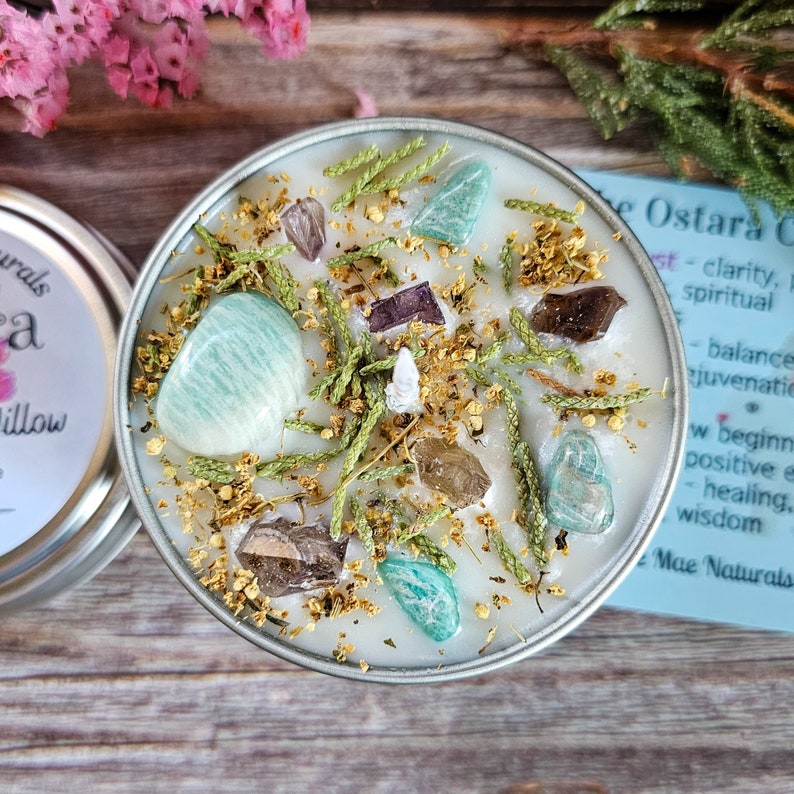 Peut inclure: Une bougie blanche avec des cristaux verts et bruns et des herbes incrust&eacute;s dans la cire. La bougie est dans une bo&icirc;te en argent avec une &eacute;tiquette qui indique "Ostara Candle - clart&eacute;, &eacute;quilibre spirituel, rajeunissement, nouveaux commencements, &eacute;nergie positive, gu&eacute;rison, sagesse, Mae Naturals."