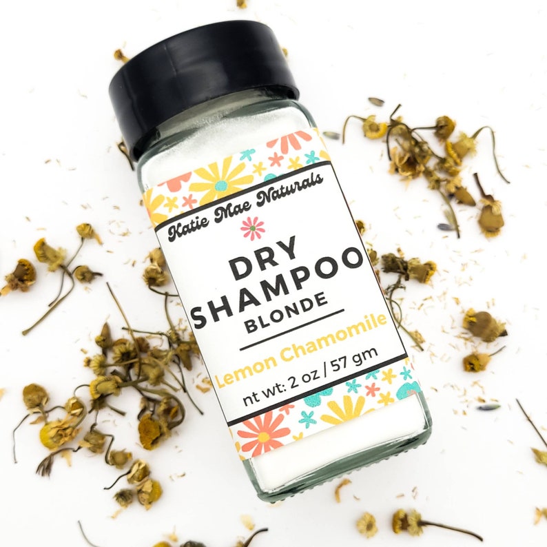 May include: A small glass jar with a white label that says "Katie Mae Naturals Dry Shampoo Blonde Lemon Chamomile nt wt: 2 oz/57 gm". The label has a floral design with pink, yellow, and blue flowers. The jar is filled with a white powder.