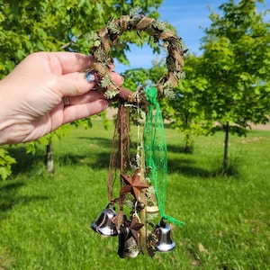 Pu&ograve; includere: Una ghirlanda verde e marrone con campane, stelle e nastri che pendono da essa. La ghirlanda &egrave; realizzata con materiali naturali e ha un aspetto rustico.