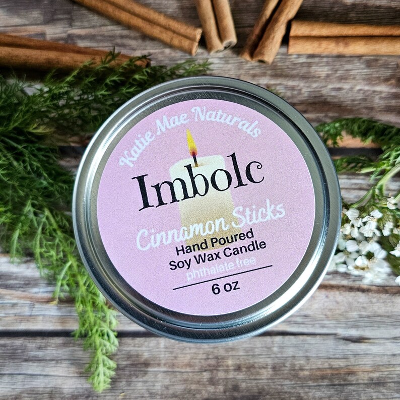 May include: A silver tin candle with a pink label that reads "Katie Mae Naturals Imbolc Cinnamon Sticks Hand Poured Soy Wax Candle Phthalate Free 6 oz".