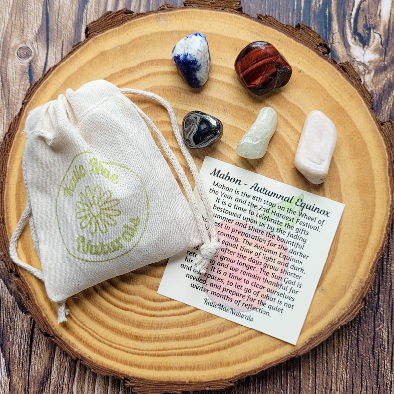 May include: A small white pouch with a green flower logo and the text "Katie Mae Naturals" sits on a wooden surface with a variety of crystals. The crystals include a blue lapis lazuli, a red tiger's eye, a black obsidian, a clear aquamarine, and a white selenite. A card with the text "Mabon - Autumnal Equinox" is also on the surface.