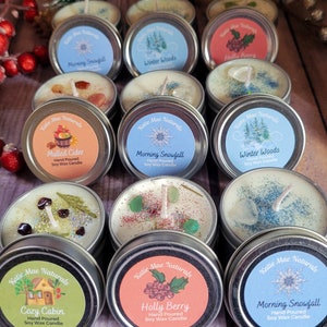 May include: Assortment of small, round, hand-poured soy wax candles in metal tins. Each tin has a different label with a winter-themed design and scent name, including "Morning Snowfall", "Winter Woods", "Mulled Cider", "Cozy Cabin", and "Holly Berry".