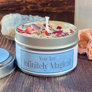 May include: A round silver metal candle tin with a white label featuring the text You Are Infinitely Magical in blue. The candle wax is topped with dried red rose petals, lavender buds, a purple crystal, and a yellow stone with a central wick.