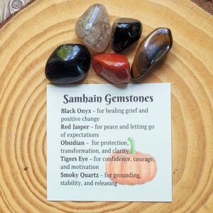 May include: A set of six polished gemstones on a wooden surface with a card that reads "Samhain Gemstones" and lists the names and properties of each stone: Black Onyx, Red Jasper, Obsidian, Tigers Eye, and Smoky Quartz.