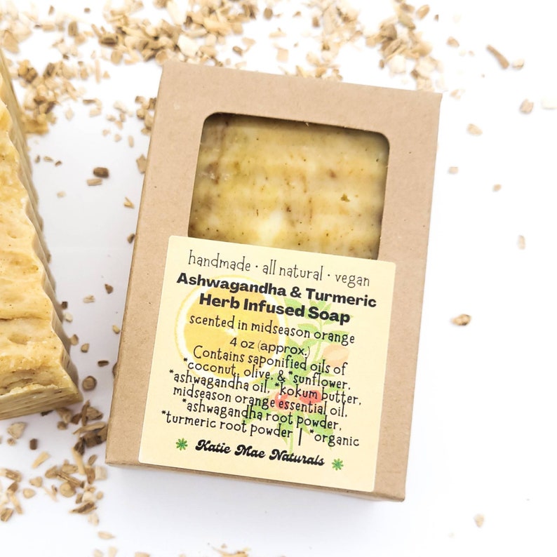 May include: A brown cardboard box with a clear window showing a bar of yellow soap. The label reads "handmade all natural vegan Ashwagandha & Turmeric Herb Infused Soap scented in midseason orange 4 oz (approx.) Contains saponified oils of coconut, olive, & sunflower. ashwagandha oil, kokum butter, midseason orange essential oil, ashwagandha root powder, turmeric root powder | organic *Katie Mae Naturals*"