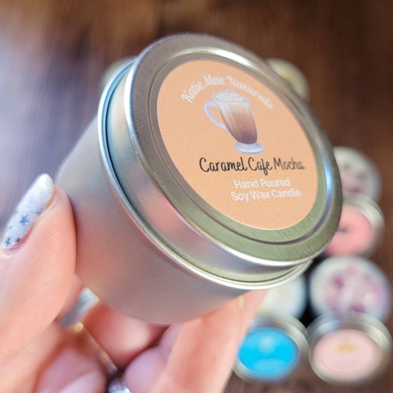 Puede incluir: Una peque&ntilde;a vela en una lata plateada con una etiqueta que dice "Katie Mae Naturals Caramel Cafe Mocha Hand Poured Soy Wax Candle". La etiqueta presenta una ilustraci&oacute;n de dibujos animados de una taza de caf&eacute;.