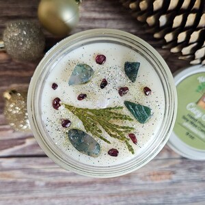 Puede incluir: Una vela de cera de soja en un tarro de vidrio transparente, decorada con piedras preciosas verdes y rojas, purpurina y una ramita de vegetaci&oacute;n. La vela tiene 7,6 cm de di&aacute;metro y tiene cera blanca. La tapa es blanca con texto verde.