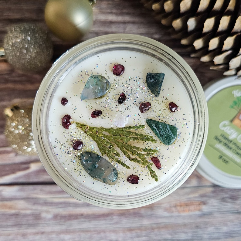Puede incluir: Una vela de cera de soja en un tarro de vidrio transparente, decorada con piedras preciosas verdes y rojas, purpurina y una ramita de vegetaci&oacute;n. La vela tiene 7,6 cm de di&aacute;metro y tiene cera blanca. La tapa es blanca con texto verde.