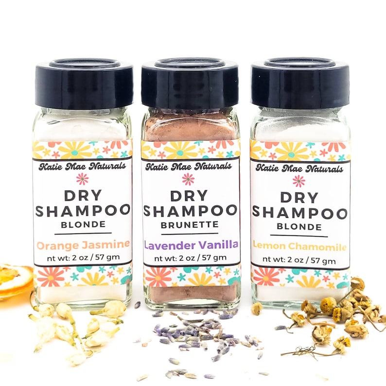 May include: Three small glass jars with black lids containing dry shampoo powder. The jars are labeled "Katie Mae Naturals Dry Shampoo" with different scents and hair colors: "Orange Jasmine Blonde", "Lavender Vanilla Brunette", and "Lemon Chamomile Blonde".