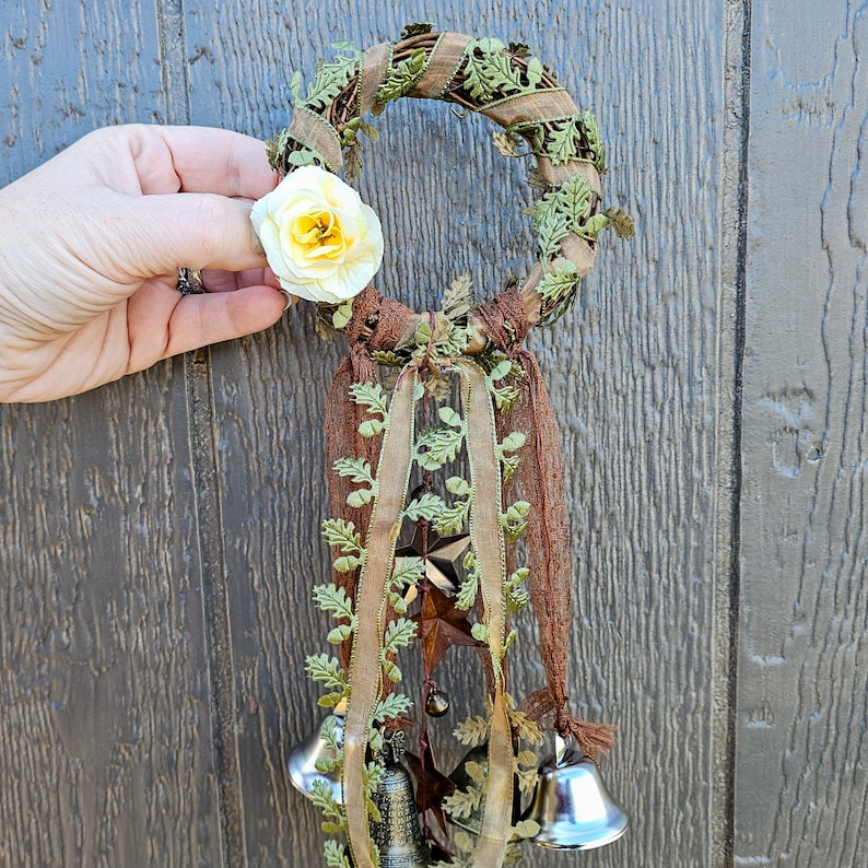 May include: A green and brown wreath with a white flower and silver bells hanging from it. The wreath is decorated with green leaves and brown ribbon.