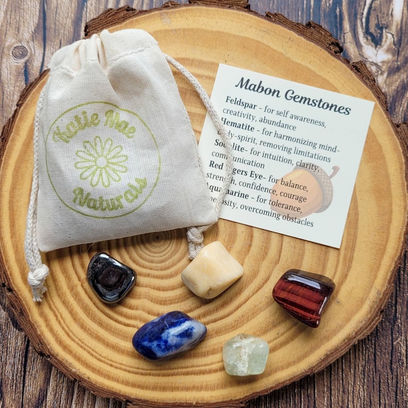 May include: A set of seven gemstones in a small pouch with a label that reads "Mabon Gemstones" and lists the names and properties of each stone. The stones are: Feldspar, Hematite, Sunstone, Red Tiger's Eye, Aquamarine, and a clear stone.