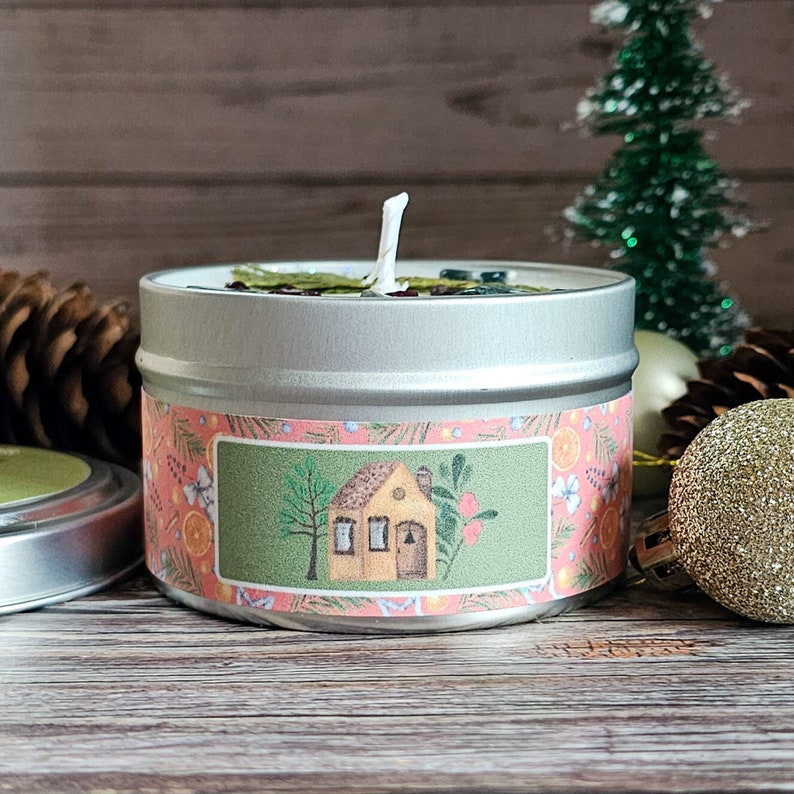 May include: A silver tin candle with a green and pink label featuring a small house illustration. The candle is surrounded by pine cones and a gold ornament.