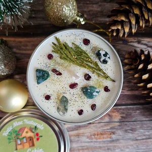 May include: A white candle with a green sprig of cedar, red and green glitter, and small green and white stones embedded in the wax. The candle is in a silver tin with a label that reads "Katie Mae Naturals Cabin".