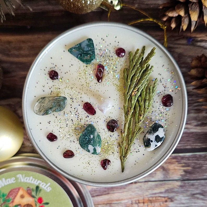 May include: A white candle with green sprigs, red and green crystals, and silver glitter. The candle is in a clear glass jar with a silver lid.