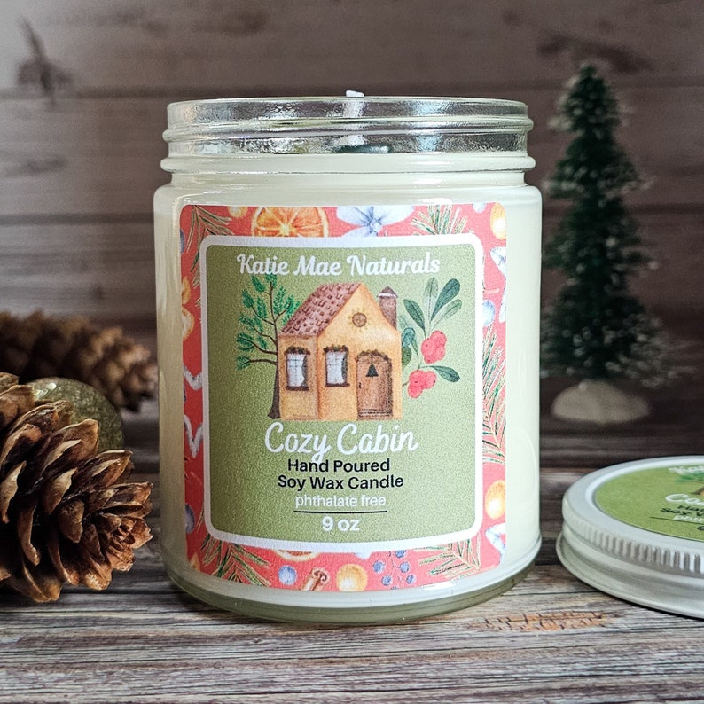 Puede incluir: Una vela en tarro de vidrio con una etiqueta que dice "Cozy Cabin". La etiqueta presenta una ilustraci&oacute;n de una casa y el texto "Hand Poured Soy Wax Candle". La vela es de 255 gramos y tiene una tapa plateada. El fondo incluye pi&ntilde;as y un peque&ntilde;o &aacute;rbol de Navidad.