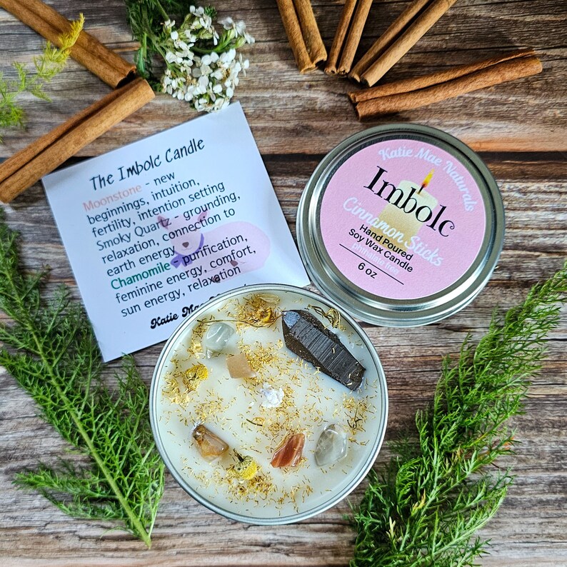 May include: A white soy wax candle with crystals and gold flakes in a silver tin. The label reads "Katie Mae Naturals Imbole Cinnamon Sticks Hand Poured Soy Wax Candle 6 oz".