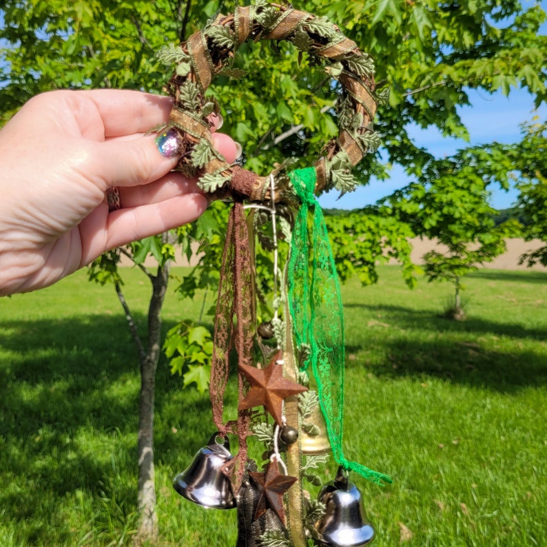 Pu&ograve; includere: Una ghirlanda di ramoscelli marroni con dettagli in pizzo e nastro verde e marrone. La ghirlanda ha cinque campane d'argento e due ciondoli a forma di stella dorata.