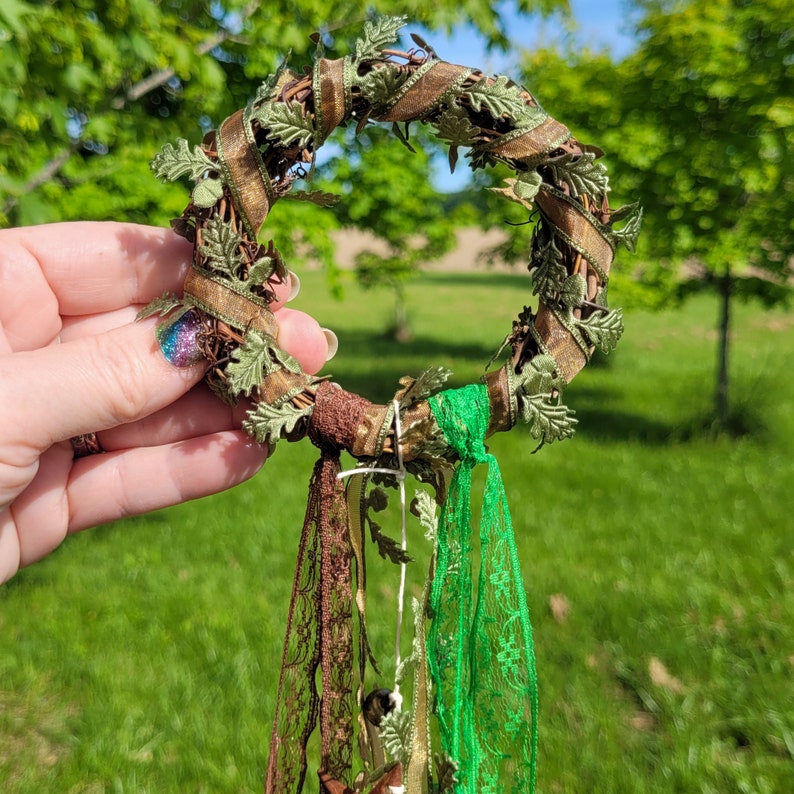 Pu&ograve; includere: Una ghirlanda verde e marrone con una variet&agrave; di foglie verdi e viti marroni. La ghirlanda &egrave; decorata con nastri verdi e marroni e ha uno spago bianco che pende dal fondo.