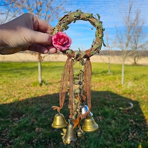 May include: A brown twig wreath with a pink fabric flower and multiple gold bells hanging from it. The wreath is decorated with green leaves and brown ribbon.
