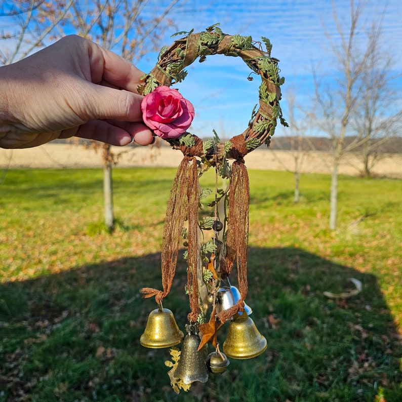May include: A brown twig wreath with a pink fabric flower and multiple gold bells hanging from it. The wreath is decorated with green leaves and brown ribbon.